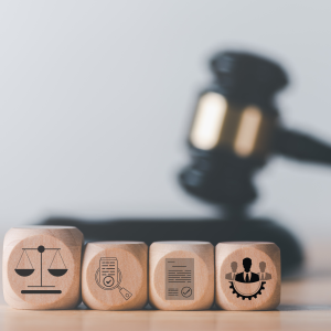 In the foreground are four wooden blocks with simple icons on each. A set of scales, a magnifying glass over a document, a letter with a seal affixed, and a group of three men. In the background is a blurred judge's gavel resting on the gavel block